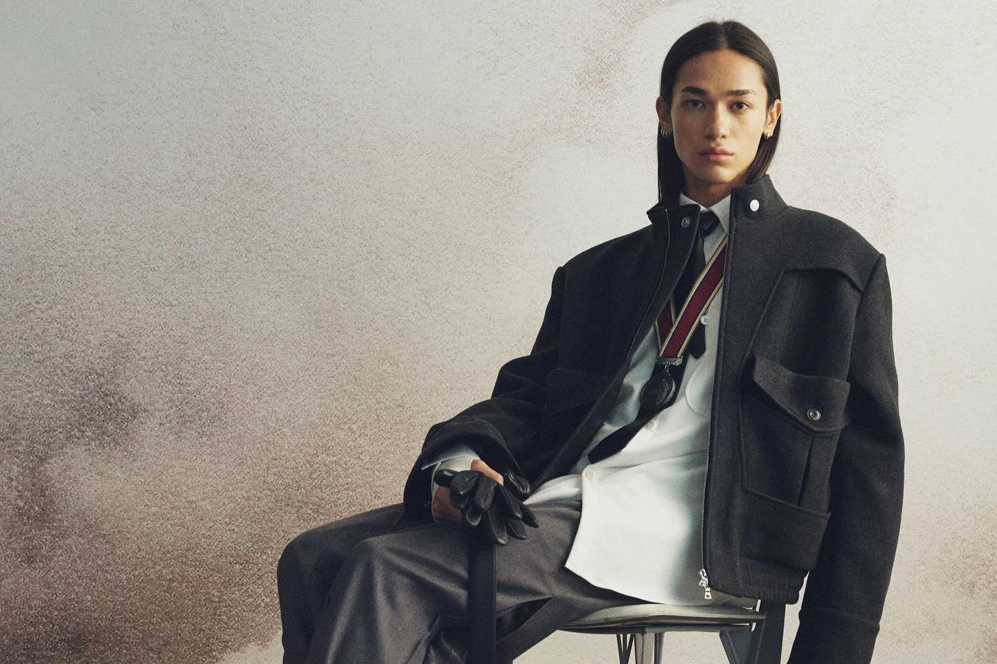 Nullus sitting on a chair wearing a black jacket and gray pants against a neutral background